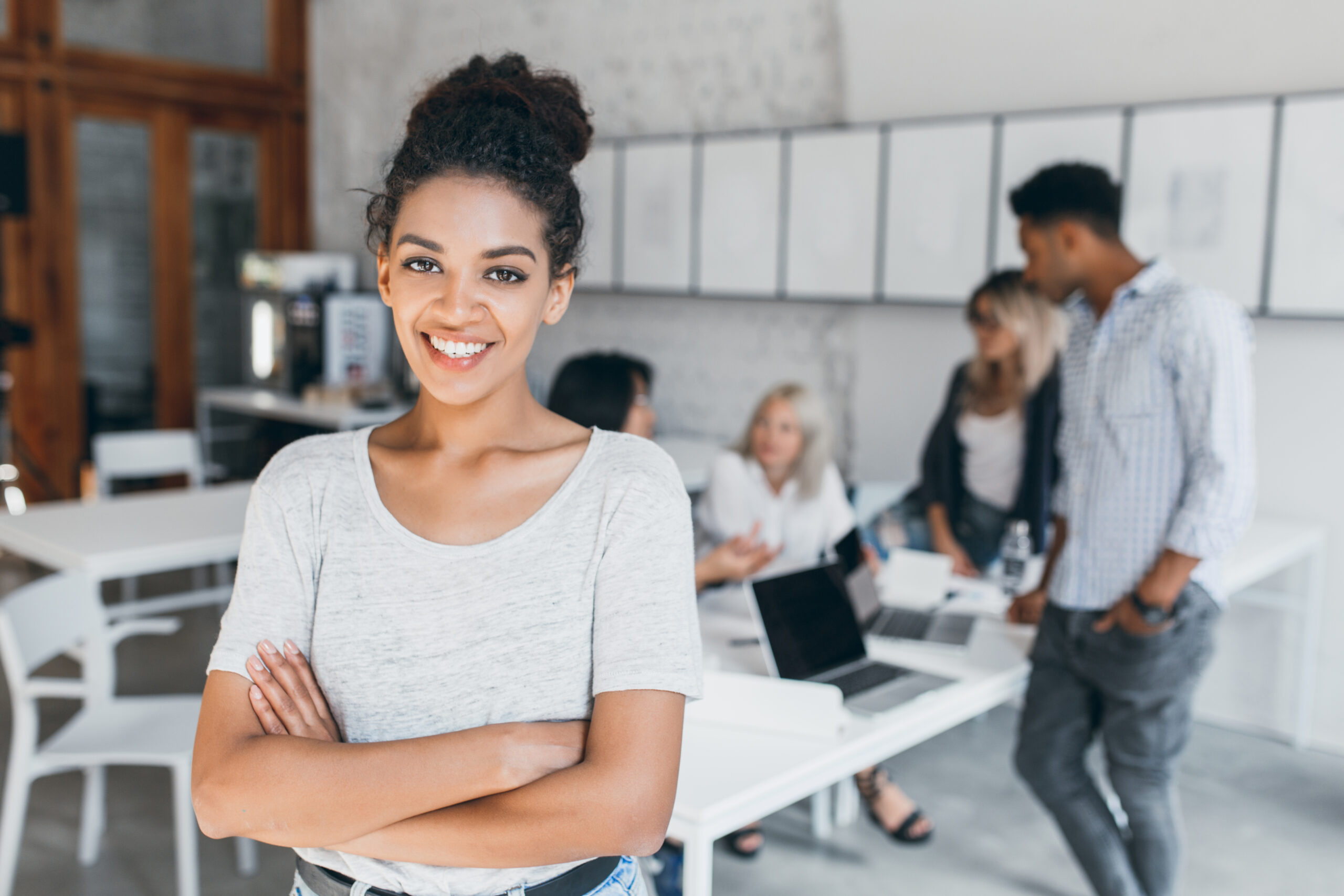 pleased woman with light brown skin posing with crossed arms and smiling, while people behind her working. indoor portrait of tired students with laptop and african curly girl on foreground..