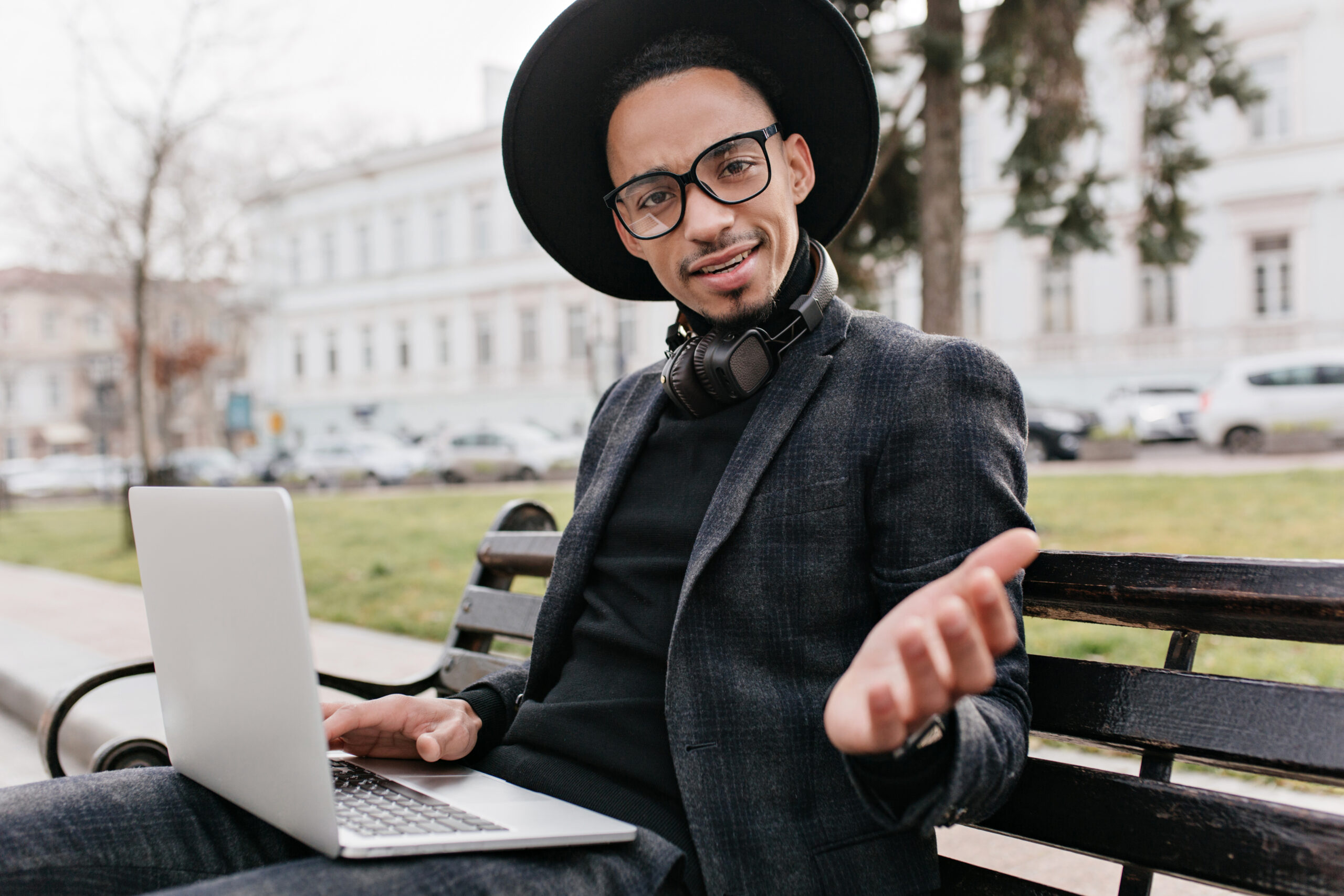 african freelancer expressing amazement while working with laptop in square. outdoor photo of surprised black man wears hat, sitting on bench and holding computer..